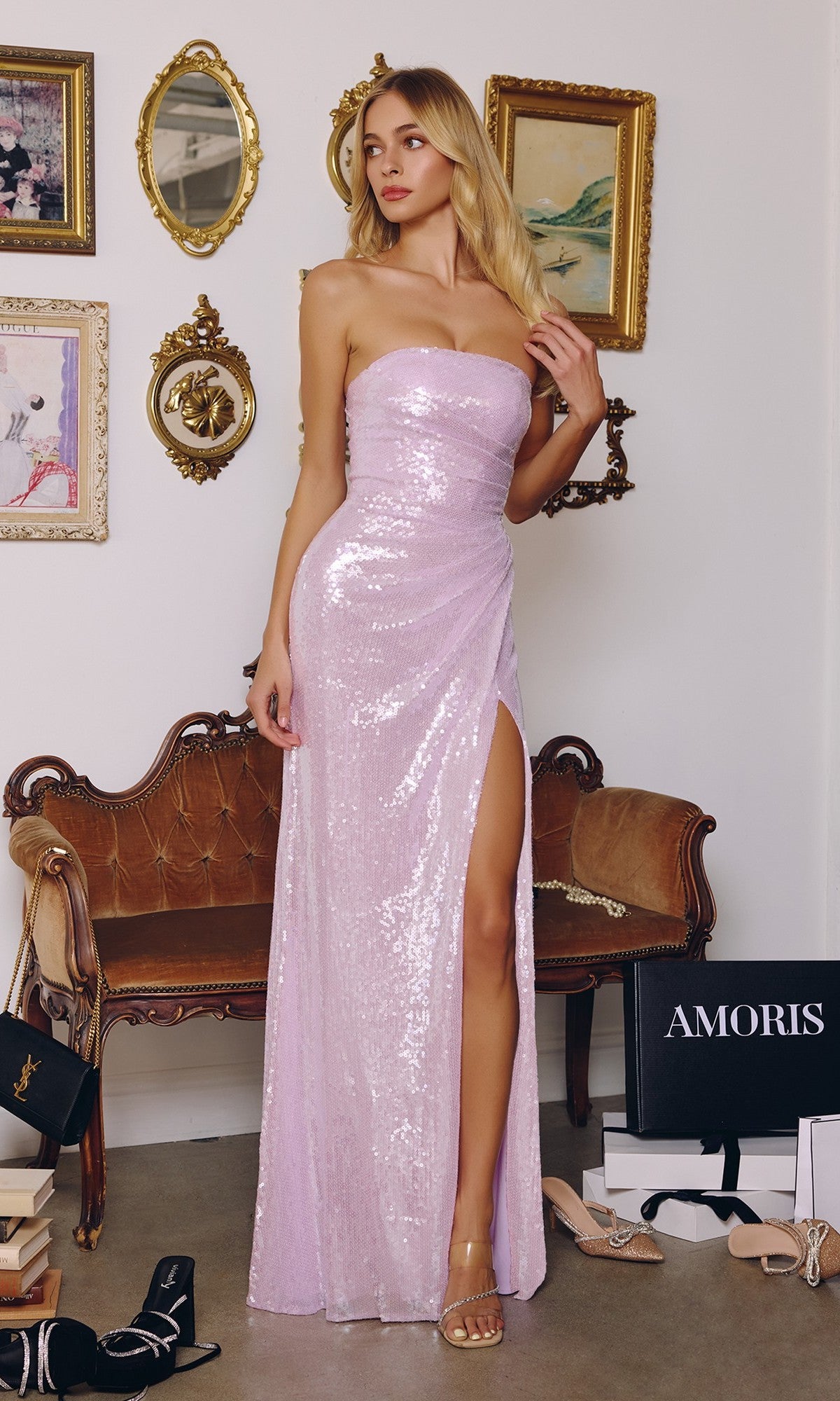 A woman in a lavender Amoris T724 long prom dress with a strapless sequin bodice and thigh-high slit stands in a decorated room filled with vintage furniture, framed art, and luxury shoe boxes.